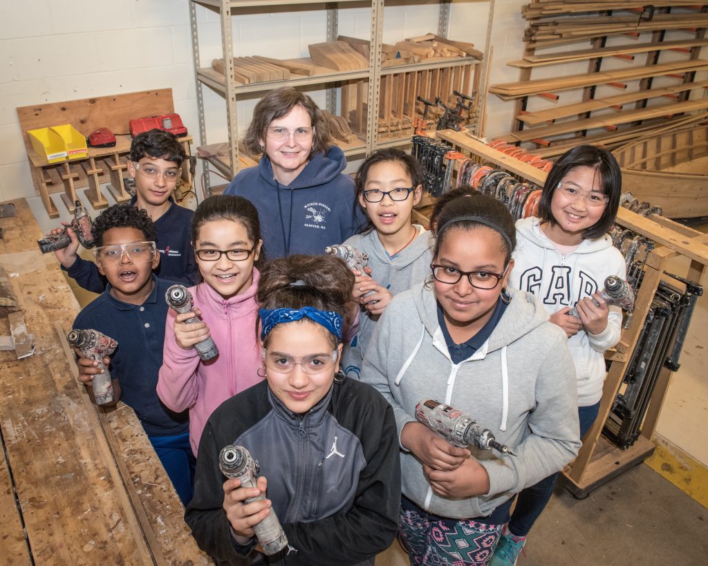 Smiling children wearing protective goggles hold power tools as an instructor looks on