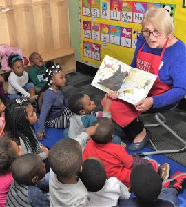 Children listen intently as a blond woman reads a storybook to them