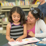 Smiling young girl reads to encouraging mother in library