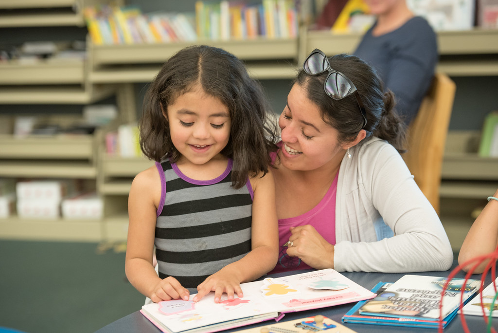 Smiling young girl reads to encouraging mother in library