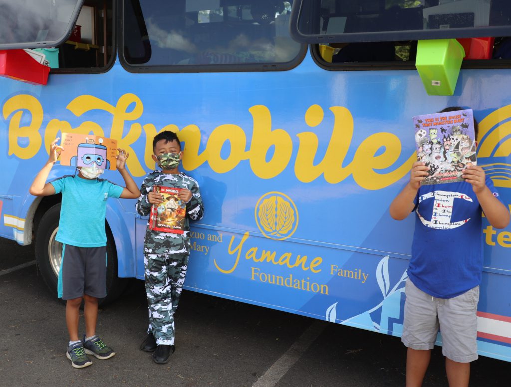 three boys hold up books in front of the Hawaii Literacy bookmobile