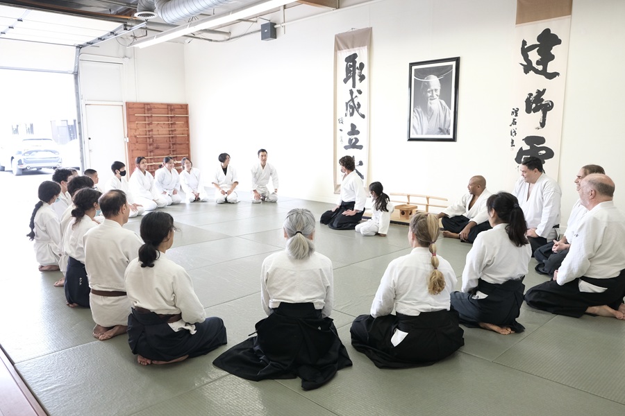 Students in white martial arts uniforms sit in a circle on dojo mats during a group discussion, with calligraphy banners and a portrait displayed on the wall behind them.