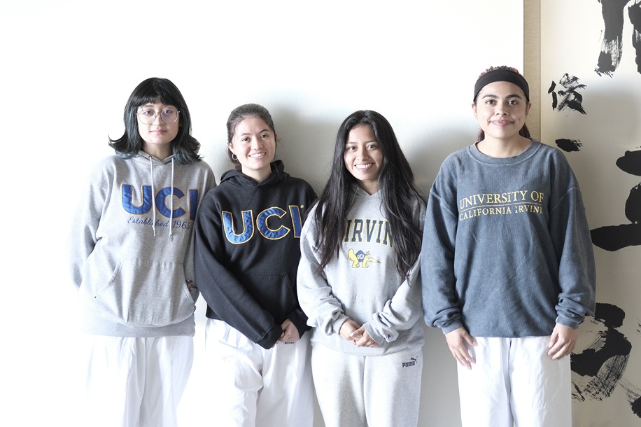 Four female students wearing University of California, Irvine sweatshirts stand together inside a dojo, smiling at the camera.