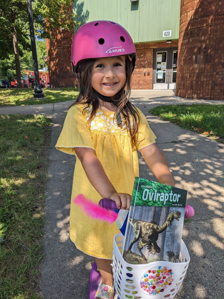 Young girl riding a scooter with a dinosaur book from The Free Book Buggie